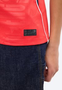 Κόκκινο αθλητικό πουκάμισο με μαύρη ετικέτα «Engineered» πάνω από σκούρο μπλε τζιν σε πλευρά και χέρι ενός ατόμου.