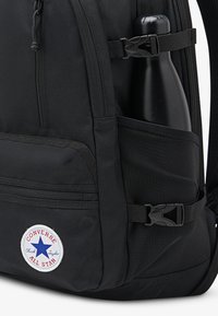 Svart ryggsekk i stoff med en synlig sidelomme som inneholder en svart vannflaske. Har en rund hvit og blå Converse-logo.