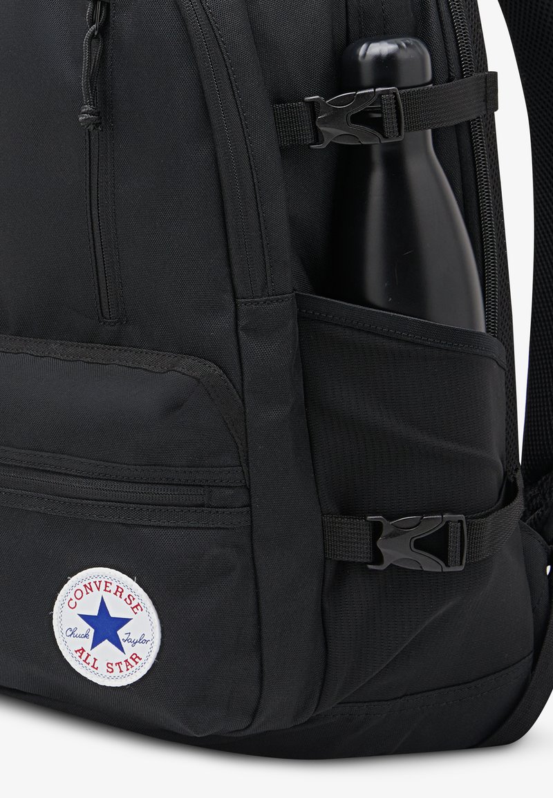 Sort stofrygsæk med en synlig sidelomme med en sort vandflaske. Indeholder et rundt hvidt og blåt Converse-logo patch.