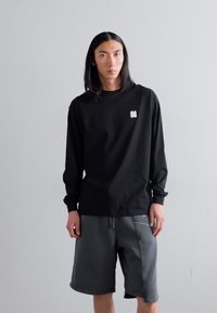 Schwarzes langärmliges T-Shirt mit einem kleinen weißen Logo auf der Brust, kombiniert mit grauen Shorts, die über ein kontrastierendes Nähdetail verfügen.