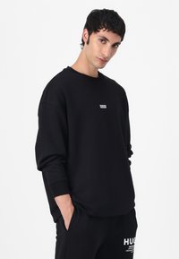 Black crew-neck sweatshirt made of smooth fabric. Features a small white logo patch on the chest and ribbed cuffs for a fitted look.