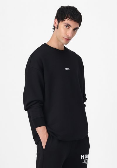 Sweatshirt noir à col rond en tissu lisse. Présente un petit patch logo blanc sur la poitrine et des poignets côtelés pour un look ajusté.
