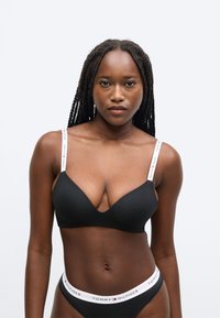 Fekete vezeték nélküli melltartó mély V alakú dekoltázzsal, sima textúrával és állítható pántokkal, melyeken fehérrel a márkanév szerepel. Hozzá illő fekete bugyi logós szalaggal.