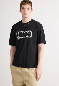 Juoda medvilninė marškinėliai su apvaliu baltu "HUGO" logotipu, trumpomis rankovėmis ir laisvu kirpimu. Sukomplektuoti su šviesiai béžiniais kelnias.