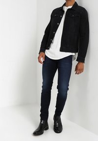 Svart denimjacka över en vit t-shirt, tillsammans med mörkblå skinny jeans och svarta chelsea boots. Neutral bakgrund.