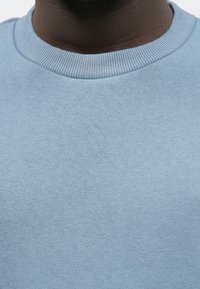 Lyseblå genser med ribbestrikket rund hals. Myk tekstur, minimal detaljer og en tettsittende silhuett. Glatt stoffutseende.