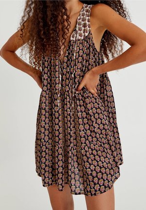 Curly-haired person wearing sleeveless black dress with multicolored floral pattern, hands on hips, against plain background.