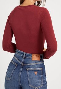 Donkerrood shirt met lange mouwen, gecombineerd met blauwe spijkerbroek. De spijkerbroek heeft een leren merkembleem en opvallende stiksels op de zakken.