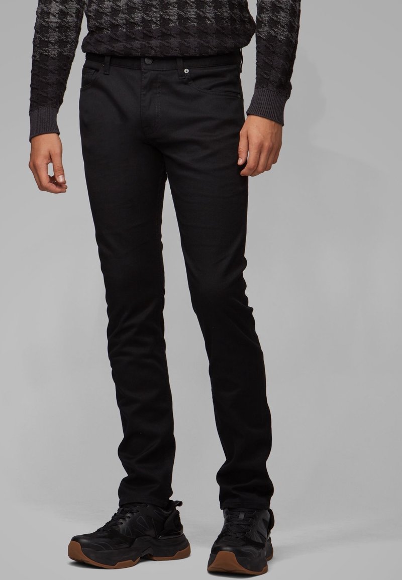 Jeans noirs slim fit avec une texture lisse, un design à cinq poches et une fermeture à bouton à l'avant, associés à des baskets foncées.