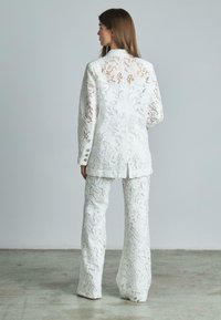 Completo bianco in pizzo con un blazer sartoriale decorato da un motivo floreale e pantaloni abbinati. Il tessuto è trasparente con dettagli intricati.