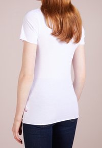 Camiseta blanca de manga corta, diseño ajustado y detalles de abertura lateral. Textura suave con un pequeño acento rojo en el dobladillo.