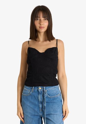 Giovane donna con capelli castani lisci che indossa un top nero senza maniche e jeans blu a vita alta, in piedi di fronte a uno sfondo bianco.