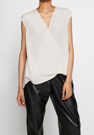 Mouwloze witte V-hals blouse gecombineerd met zwarte leren broek, gedragen door een staande persoon tegen een effen achtergrond.