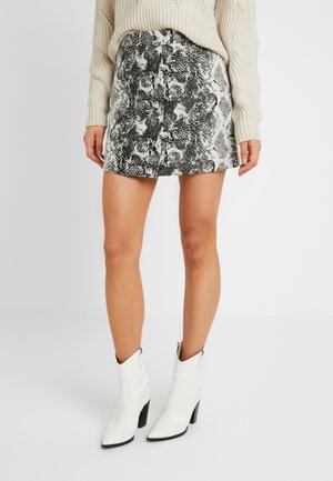 Kvinde iført en beige striktrøje, sort-hvid mininederdel med slangeskindsprint og knapper foran samt hvide ankelstøvler med hæl mod en ensfarvet baggrund.