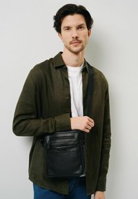 Mężczyzna w zielonej koszuli z guzikami na białym podkoszulku i niebieskich dżinsach, trzymający czarną skórzaną torbę na ramię z zamkiem, stojący na tle białej ściany.