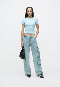 Világoskék rövid felső, laza szabású, koptatott farmer cargo nadrág zsebekkel, és fekete kézitáska. A modell fekete hegyes orrú cipőt visel.