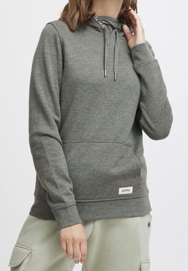 Personne portant un sweat à capuche gris avec cordons et poche avant, associée à un pantalon cargo de couleur claire, main levée près du visage.