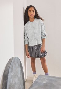 Cardigan lavorato a maglia di colore grigio chiaro con bottoni, abbinato a una gonna plissettata argentata lucida. La tenuta è completata da sneakers bianche e calzini viola.