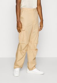 Beige cargobyxor med sidofickor, tillverkade av bomullsblandat tyg. Löst passande med rak ben-design, ihopparat med vita sneakers.