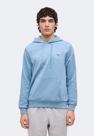 Joven con cabello rizado y oscuro que lleva una sudadera con capucha azul clara con bolsillo frontal y pantalones deportivos grises, de pie frente a un fondo simple.