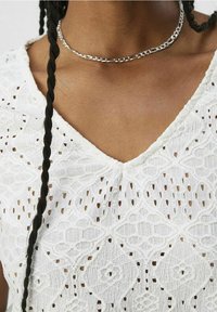 Weißes, strukturiertes Spitzentop mit V-Ausschnitt und filigranen Mustern. Eine silberne Kettenhalskette sitzt am Hals und verstärkt den Look.