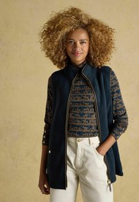 Tengerészkék bolyhos mellény arany cipzárral és részletes zsebekkel, egy mintás hosszú ujjú felsőre rétegezve. Krémszínű nadrággal párosítva.