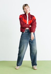 Mønstret vindjakke i mørkerød og orange med ribbede cuffs og krave; parret med højtaljede, lyse denimjeans og beige sneakers.