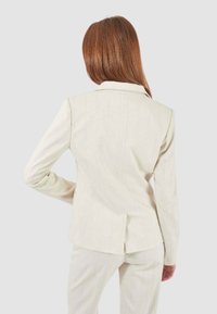 Blazer su misura beige chiaro con motivo a righe, spacco singolo sul retro e spalle strutturate. Realizzato in un tessuto liscio e testurizzato.