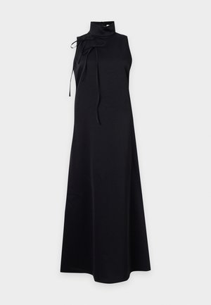 Femme aux cheveux bouclés portant une robe noire sans manches à col montant avec des liens aux épaules et des sandales noires à talons hauts et à lanières.