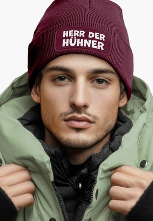 Bordeaux gestrickte Mütze mit weißem Text "HERR DER HÜHNER" und einer grünen Steppjacke über einer schwarzen Innenschicht, die einen lässigen, geschichteten Look einfängt.