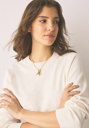 Young woman with wavy brown hair wearing a white sweater and gold charm necklace, arms crossed, looking to the side against a plain background.