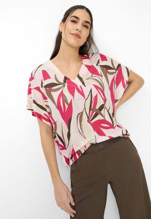 Vrouw met lang donker haar draagt een losse roze en rode bloemrijke V-hals top en een bruine broek, poseert met één hand op haar heup tegen een witte achtergrond.