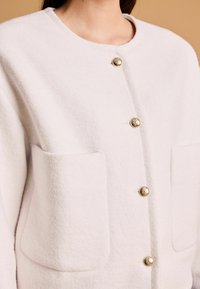 Weiße Fleecejacke mit rundem Ausschnitt, goldenen Knöpfen und zwei großen Vordertaschen. Weiche Textur mit einem klaren, modernen Design.