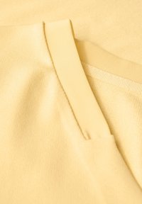 Geel stoffen kledingstuk met een gladde textuur, voorzien van een ribbelkraag en horizontale naaddetails. De close-up benadrukt de zachtheid en de kleur.
