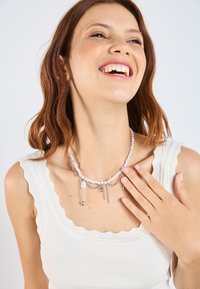 Collana bianca con perline, dal design a strati, con accenti in argento, perle e ciondoli, indossata su uno sfondo chiaro.