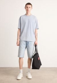 Lys blå bomuldstrøje med korte ærmer, parret med lyse denimshorts. Hvide sneakers og en sort canvas totebag med rød detalje.