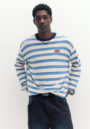 Mladý muž oblečený v modro-bielom pruhovanom tričku s dlhým rukávom a malým červeným logom na hrudi, v tmavých nohaviciach, stojaci s rukami v kapsách.