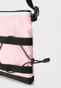 Ροζ ορθογώνια θήκη με μαύρο φερμουάρ, ρυθμιζόμενο μαύρο λουρί και ελαστικά κορδόνια στερεωμένα με μαύρες ασφάλειες σε λευκό φόντο.