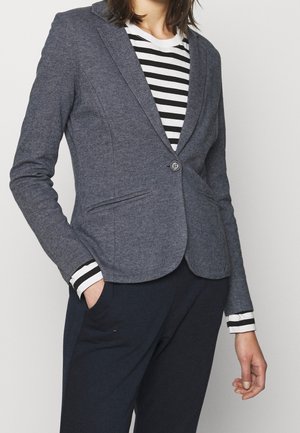 Femme portant un blazer gris boutonné par-dessus une chemise à manches longues rayée noir et blanc et un pantalon foncé, une main dans la poche.