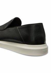 Chaussure noire en cuir à enfiler avec une finition texturée, dotée d'une semelle en caoutchouc blanc et d'un logo discret sur le talon.