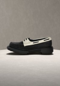 Μαύρος και λευκός δερμάτινος μοκασίνι με παχύ μαύρο τακούνι, τοποθετημένος σε ανοιχτόχρωμη επιφάνεια πάνω σε ουδέτερο φόντο.