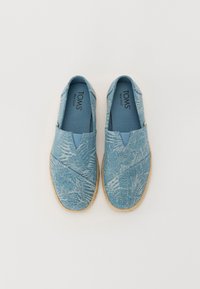 Scarpe slip-on azzurro chiaro con un tessuto testurizzato che presenta motivi di foglie di palma, una soletta imbottita e una suola naturale in juta. Marchio TOMS ben visibile.
