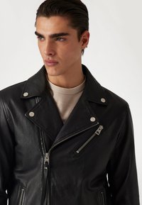 Jachetă de biker din piele neagră, cu elemente metalice argintii, buzunare cu fermoar și o cămașă tricotată texturată dedesubt, având un guler lat și un croi înclinat.