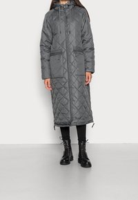 Donna che indossa un long coat imbottito grigio scuro con cappuccio, zip frontale e tasche con zip, leggings neri e stivali neri con lacci.