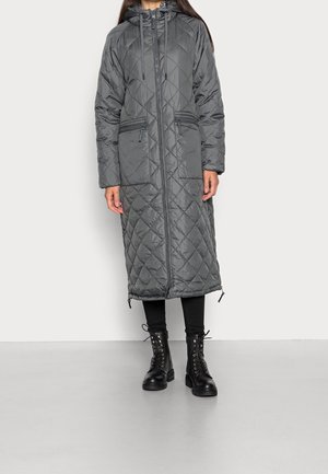 Vrouw in een lange, gewatteerde, donkergrijze gewatteerde jas met capuchon, rits aan de voorkant en ritszakken, zwarte leggings en zwarte veterschoenen.