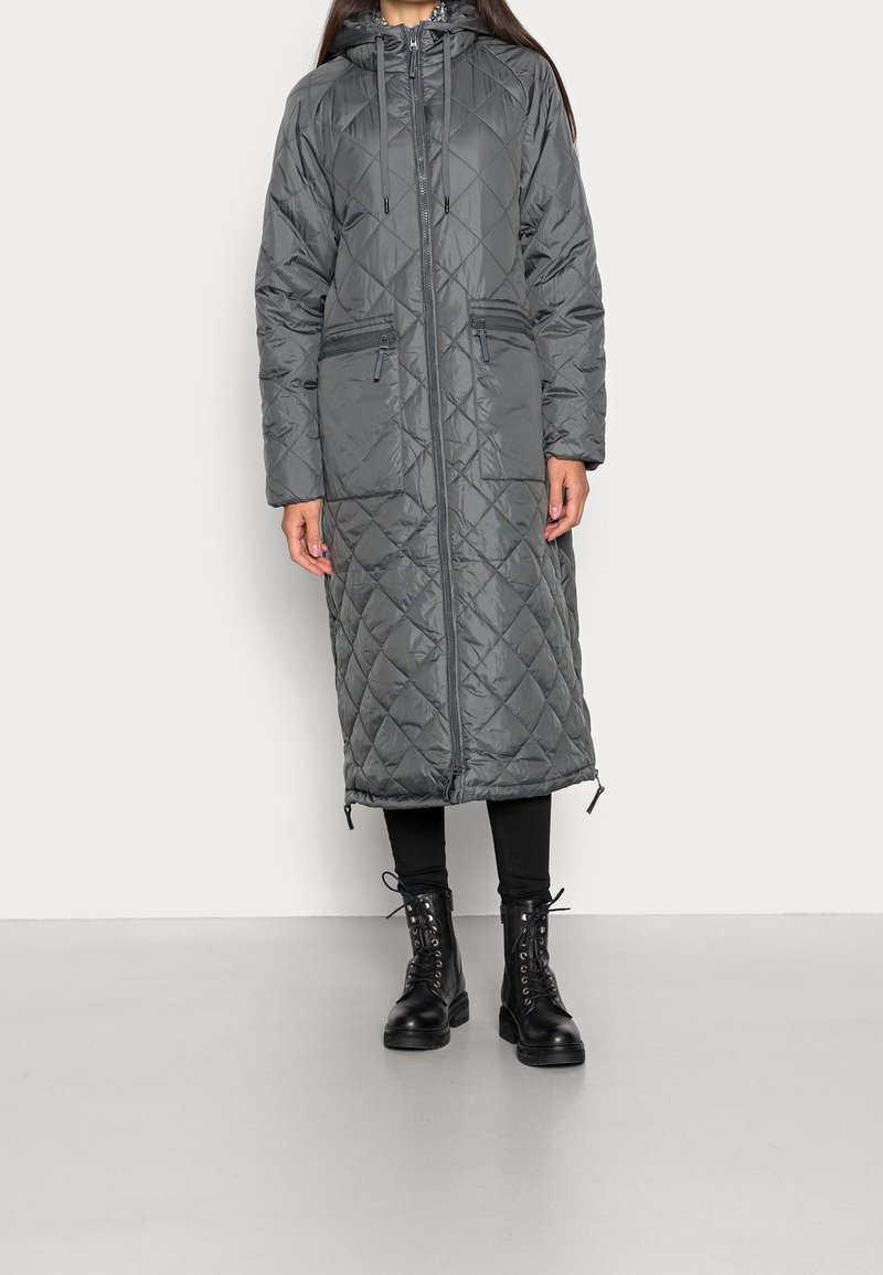 Donna che indossa un long coat imbottito grigio scuro con cappuccio, zip frontale e tasche con zip, leggings neri e stivali neri con lacci.