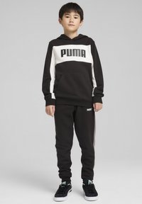 Hoodie noir et blanc avec un logo "PUMA" bien visible, associé à un pantalon de survêtement noir assorti. Le tissu semble doux avec une coupe décontractée.