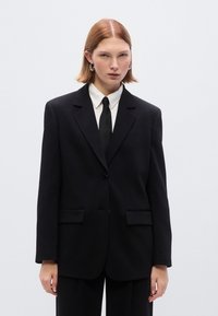 Μαύρο σακάκι με καλοραμμένη εφαρμογή, με γιακά με εγκοπές, κούμπωμα με ένα κουμπί και δύο εμπρόσθιες τσέπες. Φορεμένο πάνω από λευκό πουκάμισο και μαύρη γραβάτα.