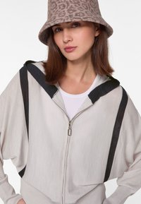 Hellgraue Zip-Hoodie mit schwarzen Akzentstreifen, weiten Ärmeln und Kapuze mit Kordelzug. Kombiniert mit einem schlichten weißen Oberteil und einem strukturierten beigen Hut.