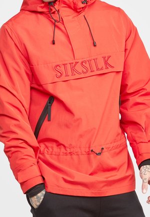 Rød hættejakke med sort lynlåslomme og "SIKSILK" branding på tværs af brystet, båret over sort tøj.